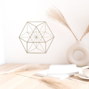 May include: A geometric design in the shape of a hexagon with a starburst pattern inside. The design is printed on a white wall. A vase with dried pampas grass sits on a wooden table with a stack of white books and a cup of coffee.