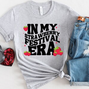 May include: A gray t-shirt with the text "In My Strawberry Festival Era" in black letters with red strawberry illustrations.