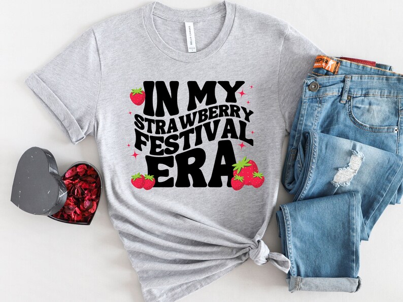 May include: A gray t-shirt with the text "In My Strawberry Festival Era" in black letters with red strawberry illustrations.