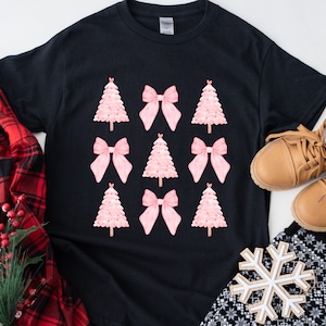 May include: Black t-shirt featuring a pattern of pink Christmas trees and bows. The shirt is surrounded by a red plaid shirt, brown boots, and a snowflake ornament, suggesting a holiday theme.