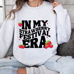 May include: A gray sweatshirt with the text "In My Strawberry Festival Era" in black with a retro font. The text is surrounded by small red strawberries.