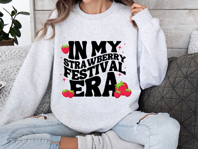 May include: A gray sweatshirt with the text "In My Strawberry Festival Era" in black with a retro font. The text is surrounded by small red strawberries.