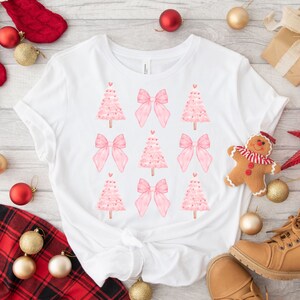 May include: White t-shirt featuring a repeating pattern of pink Christmas trees and bows. The trees have heart-shaped ornaments. The shirt is surrounded by Christmas decorations, including ornaments, a gingerbread man, and a star.
