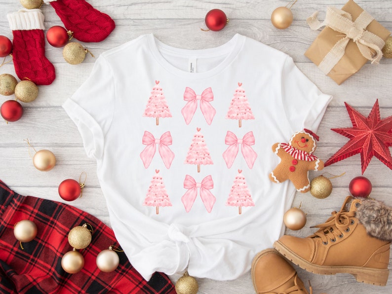 May include: White t-shirt featuring a repeating pattern of pink Christmas trees and bows. The trees have heart-shaped ornaments. The shirt is surrounded by Christmas decorations, including ornaments, a gingerbread man, and a star.