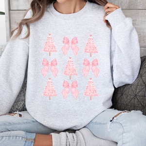 May include: A light gray sweatshirt featuring a pattern of pink Christmas trees and bows. The trees have a dotted texture and are topped with a small red heart. The bows are a solid pink color. The sweatshirt is a crew neck style.