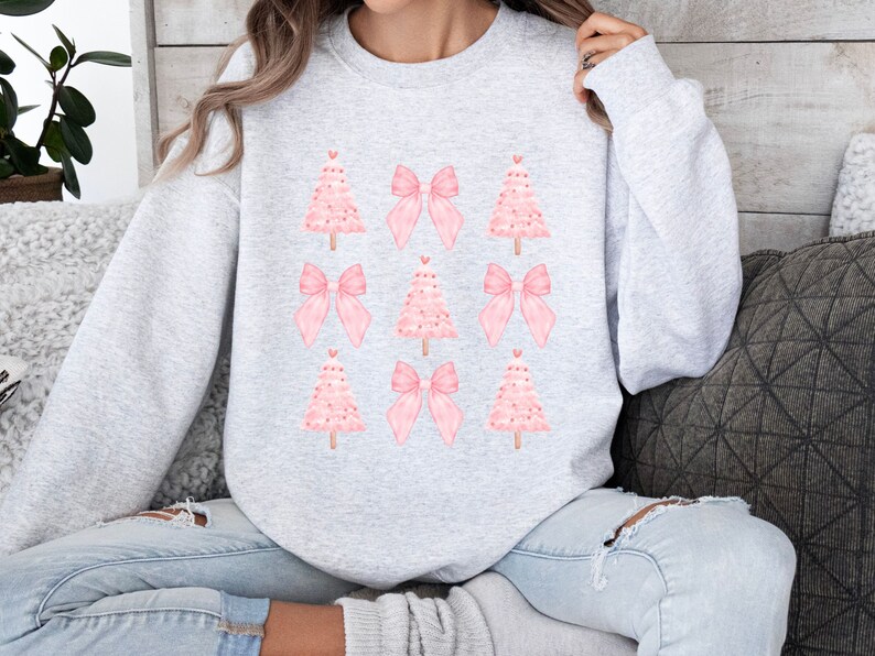 May include: A light gray sweatshirt featuring a pattern of pink Christmas trees and bows. The trees have a dotted texture and are topped with a small red heart. The bows are a solid pink color. The sweatshirt is a crew neck style.
