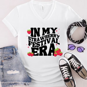 May include: White V-neck t-shirt with the text "In My Strawberry Festival Era" in black with red strawberry graphics.