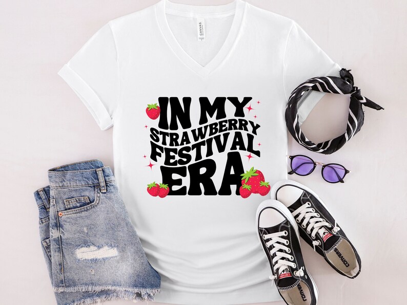May include: White V-neck t-shirt with the text "In My Strawberry Festival Era" in black with red strawberry graphics.