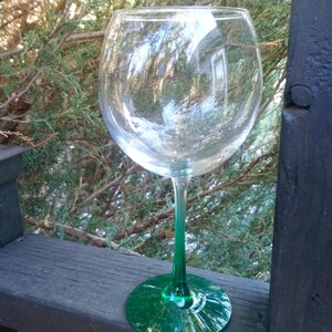 May include: A large, clear wine glass with a green stem and base. The glass is empty and reflects the surrounding environment. The stem and base are a vibrant green color. The glass is sitting on a wooden surface.