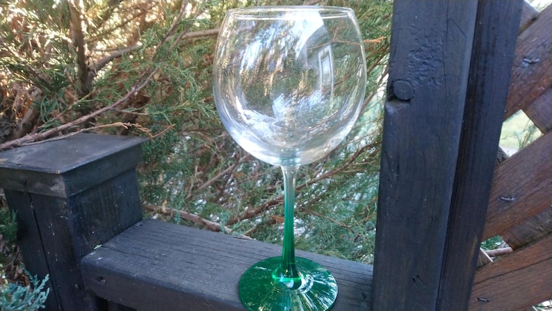 May include: A large, clear wine glass with a green stem and base. The glass is empty and reflects the surrounding environment. The stem and base are a vibrant green color. The glass is sitting on a wooden surface.