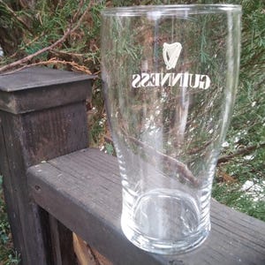 Puede incluir: Un vaso de pinta de vidrio transparente con el logotipo de Guinness impreso en el lateral. El vaso est&aacute; sobre una superficie de madera.