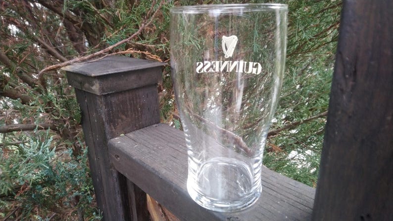 Puede incluir: Un vaso de pinta de vidrio transparente con el logotipo de Guinness impreso en el lateral. El vaso est&aacute; sobre una superficie de madera.