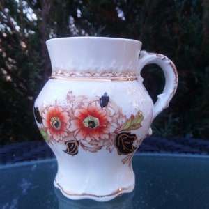 Puede incluir: Una taza de cerámica blanca con un diseño floral con flores rojas, naranjas y negras. La taza tiene un borde dorado alrededor de la parte superior y un asa.