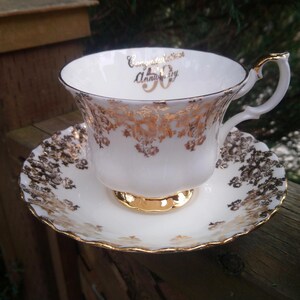 May include: A white teacup and saucer with gold trim and a floral pattern. The teacup has the words "Congratulations" and "Anniversary" written on it, along with the number "90".