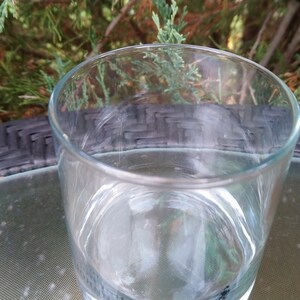 K&ouml;nnte beinhalten: Ein klarer Glasbecher steht auf einer schwarzen und grauen gewebten Oberfl&auml;che. Das Glas ist leer und scheint aus dickem Glas zu sein.