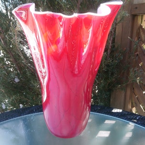 May include: A red glass vase with a ruffled top. The vase is sitting on a table.