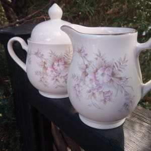 May include: A white ceramic creamer and sugar set with a floral design. The creamer has a gold rim and the sugar bowl has a lid.