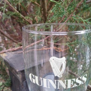 Puede incluir: Un vaso de pinta de vidrio transparente con el logotipo de Guinness y un dise&ntilde;o de arpa impreso en la parte delantera.
