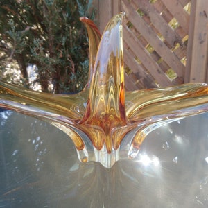 May include: A large amber glass bowl with a unique, abstract design. The bowl has a starburst shape with long, thin arms that curve upwards.