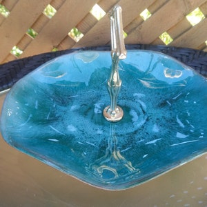 Puede incluir: Un bol de servir de vidrio azul turquesa con un asa de metal plateado. El bol tiene una forma ondulada, orgánica y un acabado brillante.