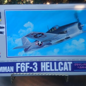 May include: A model airplane kit of a Grumman F6F-3 Hellcat in 1/48 scale. The box art features a blue sky with clouds and a silver airplane with a red, white, and blue star on the wing.