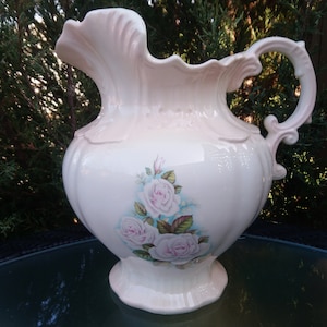 May include: A large, white ceramic pitcher with a pink glaze and floral design. The pitcher has a wide, round body and a delicate handle. The floral design features pink roses with green leaves.