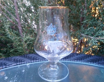 Op de afbeelding: Een helder glazen bierglas met een gouden rand en een blauw en wit logo met de tekst "Edel-Pils".