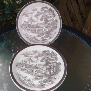 Può includere: Due piatti in ceramica bianca con bordo marrone e un design paesaggistico in bianco e nero. I piatti presentano una rappresentazione stilizzata di un ponte, alberi ed edifici.