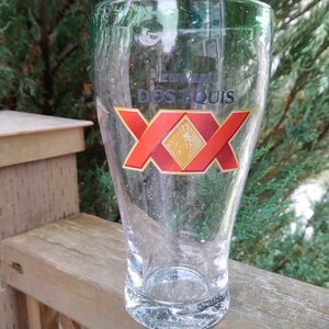 May include: A clear glass beer mug with the Dos Equis logo printed on the front. The logo is red and gold with the text "Cerveza Dos Equis" and "XX" in a large font.