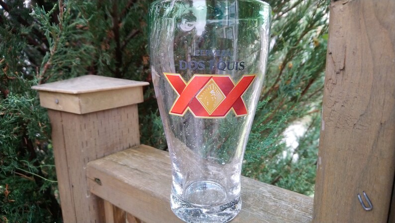 May include: A clear glass beer mug with the Dos Equis logo printed on the front. The logo is red and gold with the text "Cerveza Dos Equis" and "XX" in a large font.