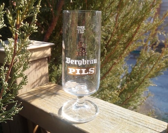 Op de afbeelding: Een helder glazen bierglas met een gouden logo met de tekst "Bergbräu Pils".