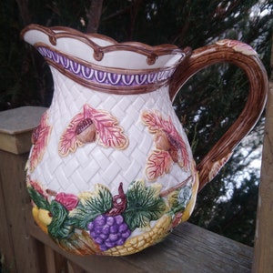 Puede incluir: Jarra decorativa de cerámica con un patrón blanco tejido, adornada con hojas de otoño, bellotas y uvas. El asa es marrón y el borde presenta un diseño morado y marrón. Esta jarra es adecuada para servir bebidas.