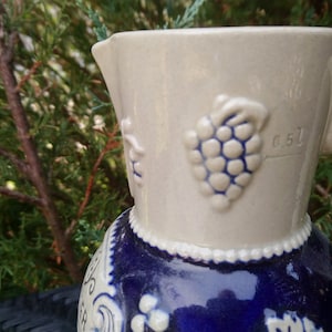 Puede incluir: Una jarra de cer&aacute;mica blanca con un dise&ntilde;o azul y blanco. La jarra tiene un asa y un pico. El dise&ntilde;o presenta uvas y un patr&oacute;n floral. La jarra tiene el n&uacute;mero "0.5 L" impreso en el lateral.