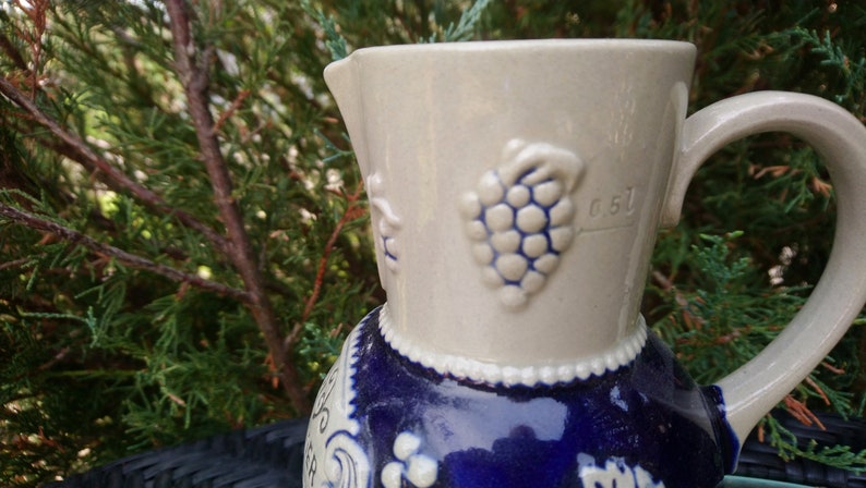 Puede incluir: Una jarra de cer&aacute;mica blanca con un dise&ntilde;o azul y blanco. La jarra tiene un asa y un pico. El dise&ntilde;o presenta uvas y un patr&oacute;n floral. La jarra tiene el n&uacute;mero "0.5 L" impreso en el lateral.