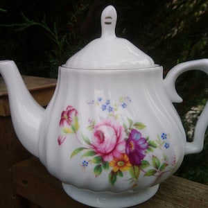 以下が含まれることがあります： ピンク、紫、黄色の花が描かれた白い磁器製のティーポット。ティーポットには蓋と取っ手があります。