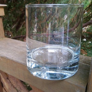 K&ouml;nnte beinhalten: Ein klares Glas mit abgerundetem Boden und geraden Seiten. Das Glas ist leer und steht auf einer Holzoberfl&auml;che.