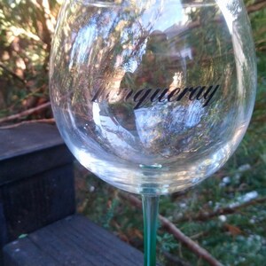 May include: A clear, stemmed wine glass with the word "Tanqueray" printed on the bowl. The glass has a green stem and is filled with a clear liquid. The background features greenery and a wooden structure.