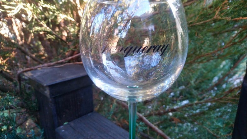 May include: A clear, stemmed wine glass with the word "Tanqueray" printed on the bowl. The glass has a green stem and is filled with a clear liquid. The background features greenery and a wooden structure.