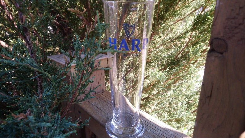 Może przedstawiać: Szklany kufel do piwa z przezroczystego szkła z nadrukowanym logo Harp Lager. Kufel stoi na drewnianej powierzchni.