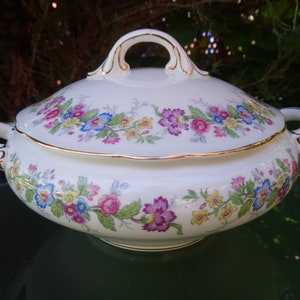 May include: A white porcelain serving dish with a floral design in shades of pink, yellow, blue, and green. The dish has a gold trim around the edge and a lid with a handle.