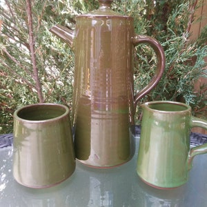 Pode incluir: Um conjunto de três canecas de cerâmica verde, incluindo um jarro com alça. As canecas são todas de um tom semelhante de verde com uma borda marrom.