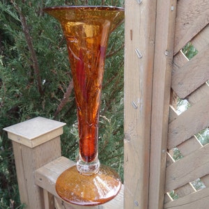 Puede incluir: Un jarrón alto de vidrio ámbar con una parte superior acampanada y una superficie texturizada. El jarrón tiene una base de vidrio transparente.