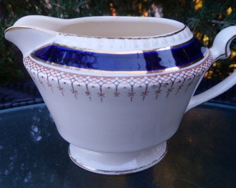 Könnte beinhalten: Weiße Keramik-Milchkanne mit einem blauen und goldenen Band um die Oberseite. Die Milchkanne hat ein zartes, filigranes Design in Braun.