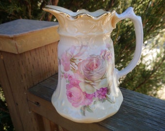 May include: A white ceramic jug with a gold rim and handle. The jug is decorated with a floral pattern of pink and white roses.