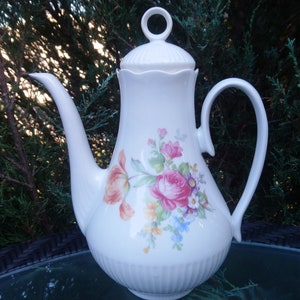 Puede incluir: Cafetera de porcelana blanca con tapa y asa curva. La cafetera está decorada con un estampado floral con flores rosas, rojas y azules. Tiene un pico largo y un anillo en la tapa.