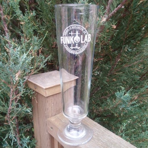 Puede incluir: Un vaso de cerveza alto y transparente de vidrio con un logotipo blanco que dice "FUNK LAB" en un círculo con una imagen estilizada de un matraz y espadas cruzadas.