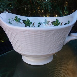May include: White ceramic gravy boat with a woven pattern and a green and white floral design around the rim.