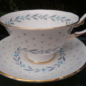 Pode incluir: Uma chávena de chá e um pires brancos com um delicado padrão floral azul e cinza. A chávena e o pires têm uma borda dourada.