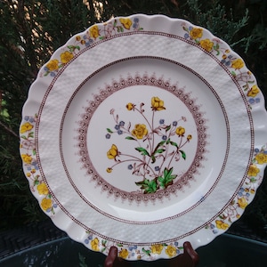 Op de afbeelding: Een wit keramiek dinerbord met een gegolfde rand en een bloemenontwerp. Het bord heeft een centraal motief van gele bloemen met groene bladeren en paarse accenten, omringd door een bruine en witte rand.
