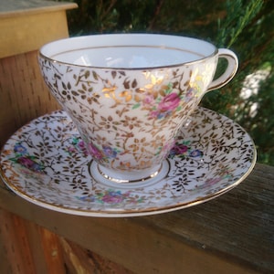 Puede incluir: Una taza de té y un platillo blancos con un patrón floral y dorado. La taza y el platillo están decorados con flores rosas, azules y amarillas.